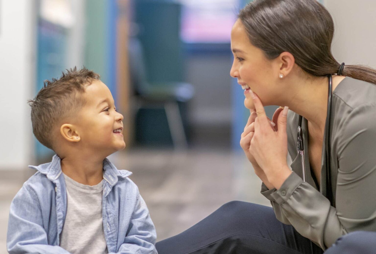 A small child is smiling up at a medical practitioner who is making a funny face at him and holding the corners of her mouth up while smiling. 