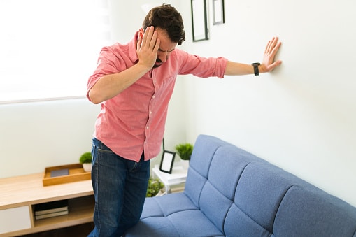 Man Experiencing Dizzy Spell Man leaning against wall in his living room, holding his head with one hand while his other is outstretched against the wall for support